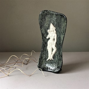 May include: A ceramic wall sculpture with a green and black textured surface. A white ceramic figure of a woman is embossed on the surface.