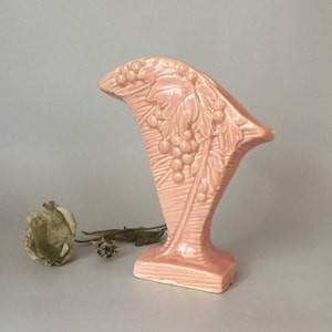 May include: A pink ceramic vase with a grapevine design. The vase is shaped like a fan and has a textured surface.