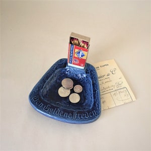 May include: Blue ceramic ashtray with the text "Den Goldene Frieden" and a small box of matches with the text "Strike on Box Matches" inside. There are four silver coins in the ashtray.