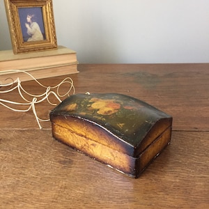 May include: A small, wooden box with a curved lid. The box is painted with a floral design in shades of yellow, orange, and brown. The box is sitting on a wooden surface.