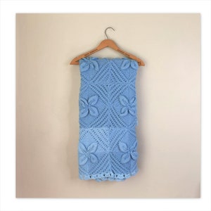 May include: A light blue crocheted blanket with a floral pattern hangs on a wooden hanger. The hanger has the words "Hotel Syracuse" and "Envoy House" printed on it.