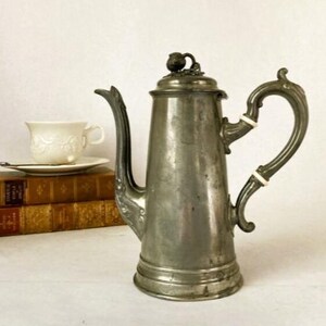 May include: A vintage silver-toned coffee pot with an ornate handle and spout. The pot has a tall, slender body and a decorative lid. A white teacup and saucer sit on a stack of old books, suggesting a classic aesthetic.