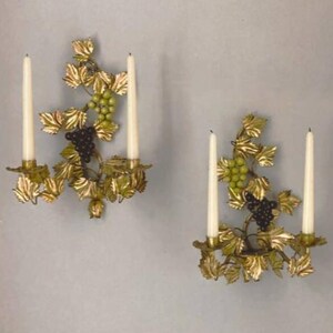 May include: Two wall sconces with a grape vine design. Each sconce features a gold-colored metal frame with leaves, green and black grapes, and two white candles. The sconces are mounted on a light gray wall.