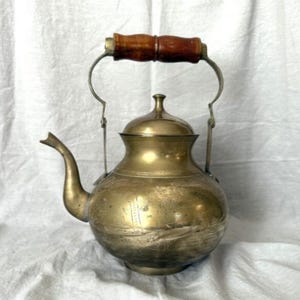 May include: Vintage brass teapot with a wooden handle and a curved spout. The teapot has a rounded body and a lid with a small knob. The handle is attached to the body with metal brackets. The teapot is likely used for brewing tea.