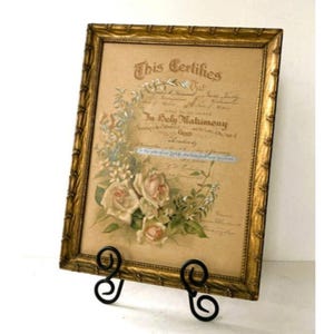 Puede incluir: Un certificado enmarcado con un marco dorado ornamentado, exhibido en un soporte de metal negro. El certificado tiene ilustraciones florales y el texto "This Certifies" y "In Holy Matrimony". El marco es rectangular con un diseño texturizado.
