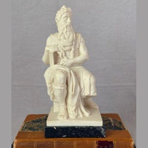 May include: A cream-colored statue of a seated figure with a long beard and draped clothing. The statue rests on a black base atop a stack of antique books with a leather cover. The figure holds a tablet.