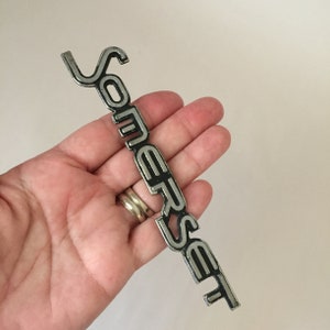以下が含まれることがあります： スタイリッシュなフォントで「サマセット」と書かれた銀と黒の金属製のエンブレムです。