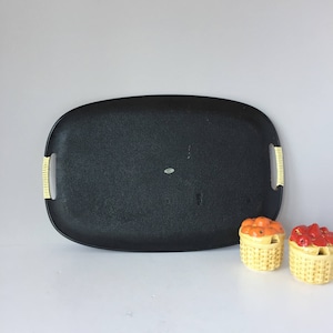 K&ouml;nnte beinhalten: Ein schwarzes ovales Serviertablett mit gelben Griffen. Das Tablett besteht aus einem strukturierten Material. Zwei kleine Keramikk&ouml;rbe mit Fruchtmotiven befinden sich im Vordergrund.