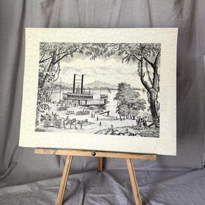 May include: Black and white print of a steamboat on a river, surrounded by trees and figures. The artwork is displayed on a small wooden easel. The steamboat has multiple decks and a tall smokestack. The scene depicts a historical river landscape.