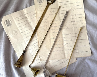 May include: An assortment of antique candle snuffers in various metallic finishes, including brass and silver, are arranged on aged paper with handwritten script. The snuffers have bell-shaped and ornate designs.