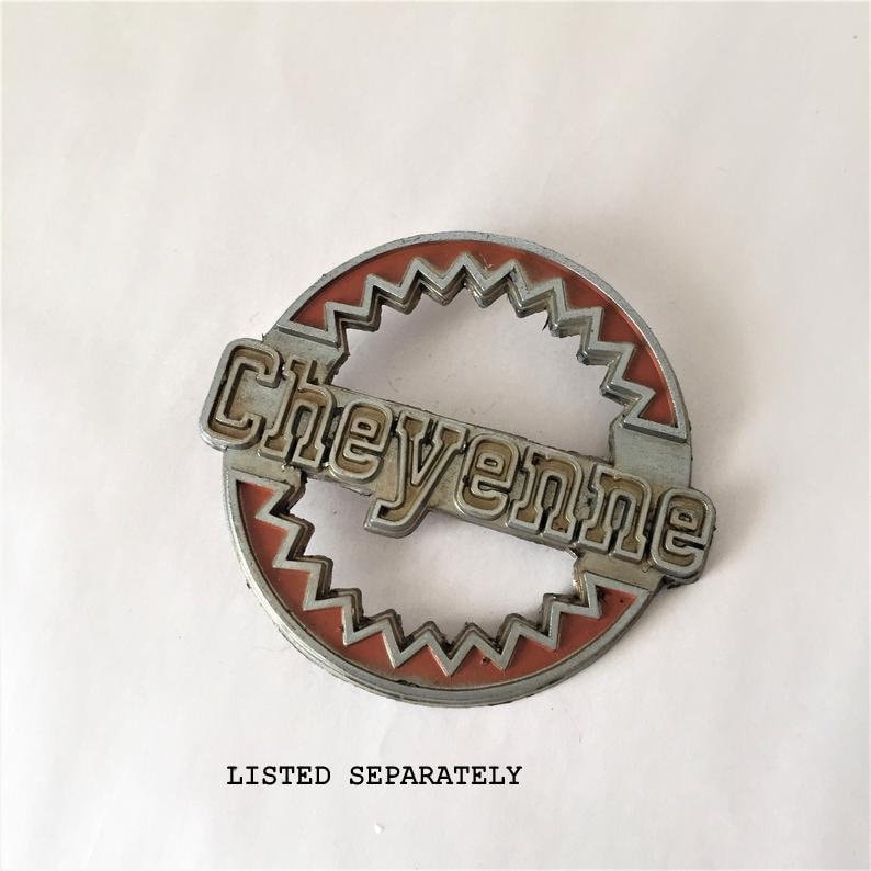May include: A silver and red metal emblem with the word "Cheyenne" in raised letters. The emblem is circular with a starburst design around the edge.