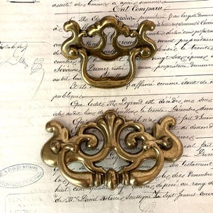 May include: Two antique brass drawer pulls with ornate scrollwork and a warm, golden finish. The pulls have a curved handle and decorative backplates, perfect for vintage furniture restoration. They rest on a backdrop of aged documents.