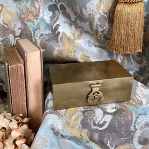 May include: A gold metal box with a decorative latch. The box is sitting on a patterned fabric with a gold tassel.