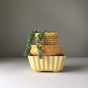 May include: A set of three yellow ceramic planters, one with a woven design, stacked on top of each other. A green succulent plant is growing out of the top planter.