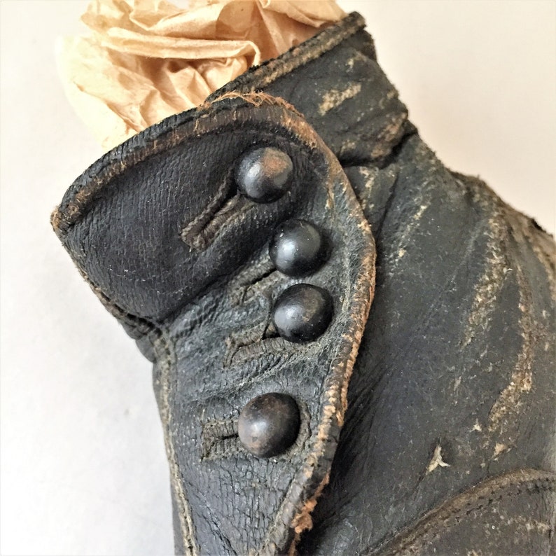 May include: Close-up of a black leather boot with four black buttons. The boot is worn and has a distressed look.