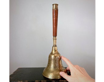 May include: A brass bell with a wooden handle. The bell has an ornate design and is sitting on a black surface.