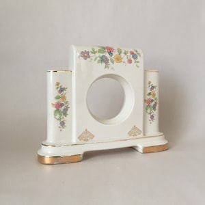 May include: White ceramic clock stand with a floral design and gold accents. The stand has a circular opening in the center for a clock face.