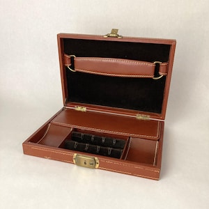 May include: Open brown leather jewelry box with a black velvet interior. The box has a handle, brass hardware, and compartments for storage. The box is open, revealing the interior and storage spaces.
