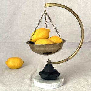 May include: A decorative brass scale with a black geometric base and a marble platform holds three yellow lemons. A single lemon rests on the off-white surface. The scale has a curved arm and a chain supporting the bowl.