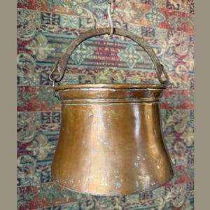 May include: A vintage copper cauldron with a curved handle, suspended from a chain. The cauldron has a wide base and tapers upwards, showing signs of age and use. The background features a patterned textile.