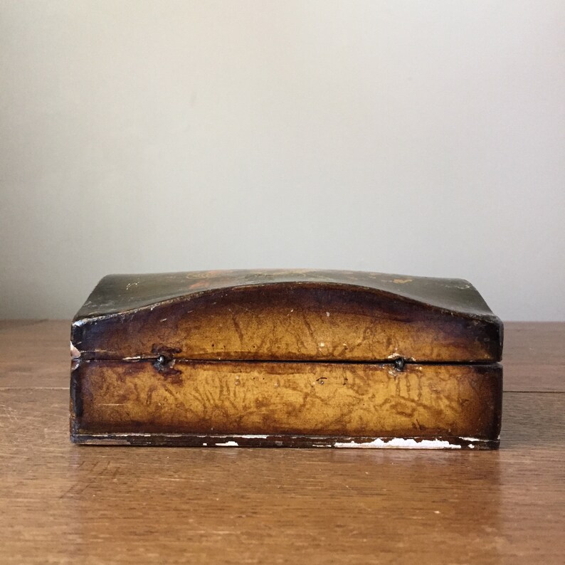 May include: A small wooden box with a curved lid. The box is painted brown with a gold-colored design. The box is closed.