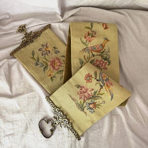 May include: A long, pale yellow needlepoint bell pull featuring floral and bird motifs in shades of pink, blue, and green. The textile is held by two ornate, antique-style brass hardware pieces with intricate scrollwork and a hanging loop.