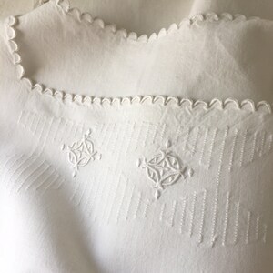 May include: White linen tablecloth with a scalloped edge and embroidered designs. The tablecloth features a repeating pattern of vertical lines and two intricate floral motifs.
