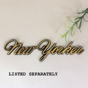 May include: Gold-colored metal emblem with the words "New Yorker" in a stylized script font. The emblem is on a white background.  LISTED SEPARATELY