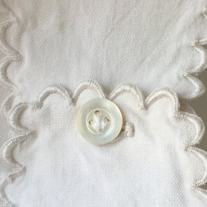May include: A white button with a scalloped edge, sewn onto a white fabric with a scalloped trim.