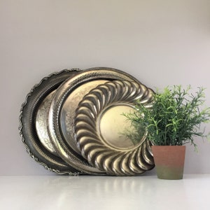 May include: A set of three silver-toned metal serving trays with a decorative design. The trays are stacked on top of each other, with the largest tray on the bottom and the smallest tray on top. A small potted plant is in front of the trays.