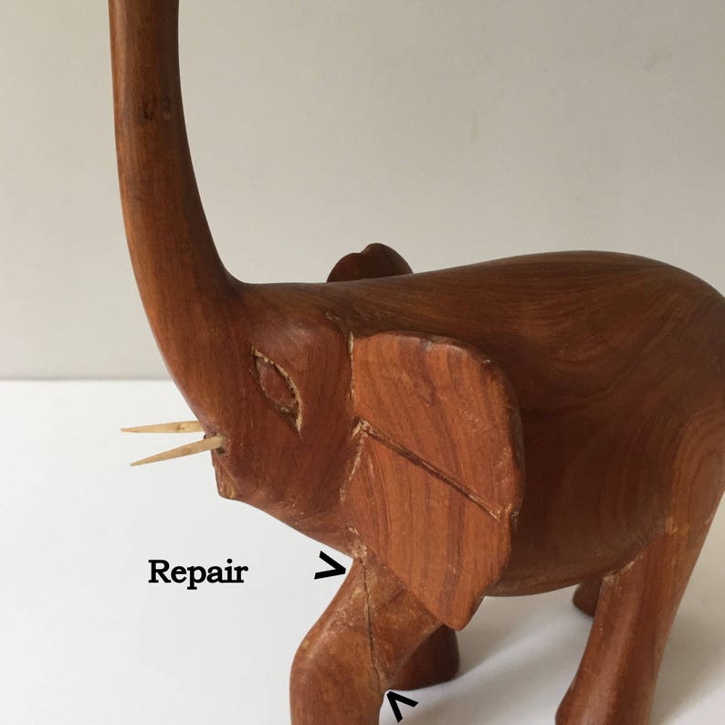 May include: A wooden elephant figurine with a long trunk and a repaired leg. The elephant is made of reddish-brown wood and has a natural finish. The figurine has a small crack in the leg, which has been repaired with a toothpick.