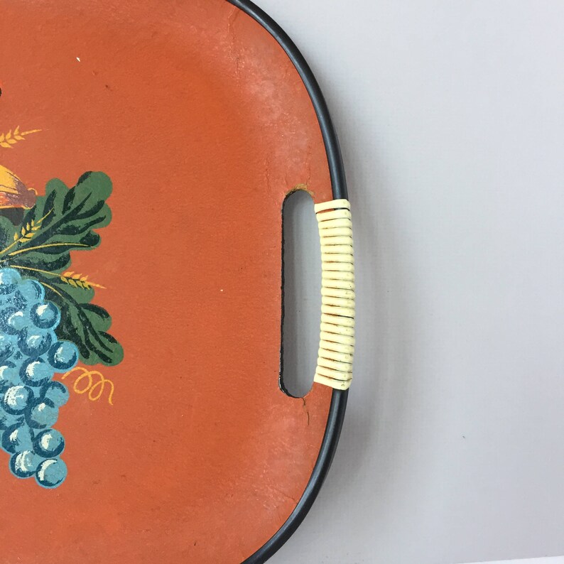 K&ouml;nnte beinhalten: Ein vintage ovales Serviertablett mit schwarzem Rand und einem geflochtenen Griff in Cremefarbe. Das Tablett ist mit einem leuchtend orangenen Hintergrund und einem Traubenranken-Design mit gr&uuml;nen Bl&auml;ttern und blauen Trauben bemalt.