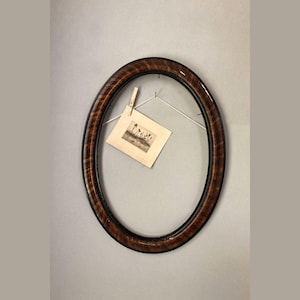 May include: An oval-shaped picture frame with a dark brown wood finish. The frame has a black inner edge and is hanging on a wall. A small photo is clipped to the frame with a clothespin.