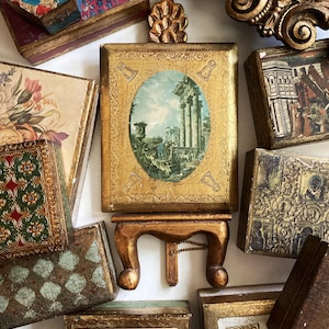 May include: An assortment of antique decorative boxes and a small wooden stand. The boxes feature various designs, including floral patterns, architectural scenes, and geometric shapes. The central piece is a gold-framed picture with an oval landscape painting.