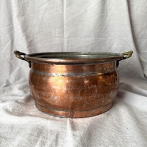 May include: A copper pot with two brass handles. The pot has a rounded body and a wide rim. The copper has a warm, reddish-brown color with some areas of tarnish. The pot is likely vintage or antique.