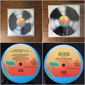 Puede incluir: Un disco de vinilo negro con un dise&ntilde;o de espiral blanco y azul. El disco est&aacute; etiquetado como "U2" y presenta los t&iacute;tulos de las canciones "New Year's Day" y "Two Hearts Beat As One".