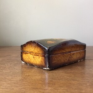 May include: A small wooden box with a hinged lid. The box is painted with a dark brown base and a gold-colored design. The box is closed and sitting on a wooden surface.