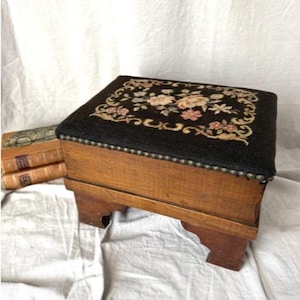 May include: An antique footstool with a dark brown, floral needlepoint top. The wooden frame is a light brown color, and the edges of the top are lined with small, silver-colored tacks. The footstool has four sturdy legs.