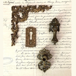 May include: Antique brass hardware pieces, including a corner bracket, keyhole cover, handle, and decorative pull, arranged on aged paper with handwritten script. The ornate designs and aged patina suggest a vintage aesthetic.