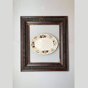 May include: An oval-shaped serving platter with a white background and a decorative border of gold, blue, and green floral designs. The platter is displayed in a dark wood frame.