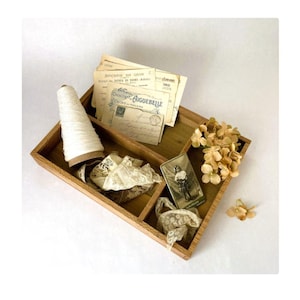 May include: A wooden tray with four compartments, filled with vintage postcards, lace, a spool of white thread, and a small card with a vintage image of a girl. The tray is decorated with dried flowers.