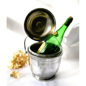 May include: A silver wine bucket with a lid, holding a green wine bottle. The bottle has a cork and a yellow label. Dried flowers are next to the bucket. The bucket is on a white surface.