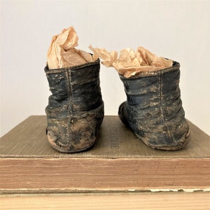 May include: A pair of black leather baby shoes with a worn and distressed look. The shoes are sitting on top of a stack of books.