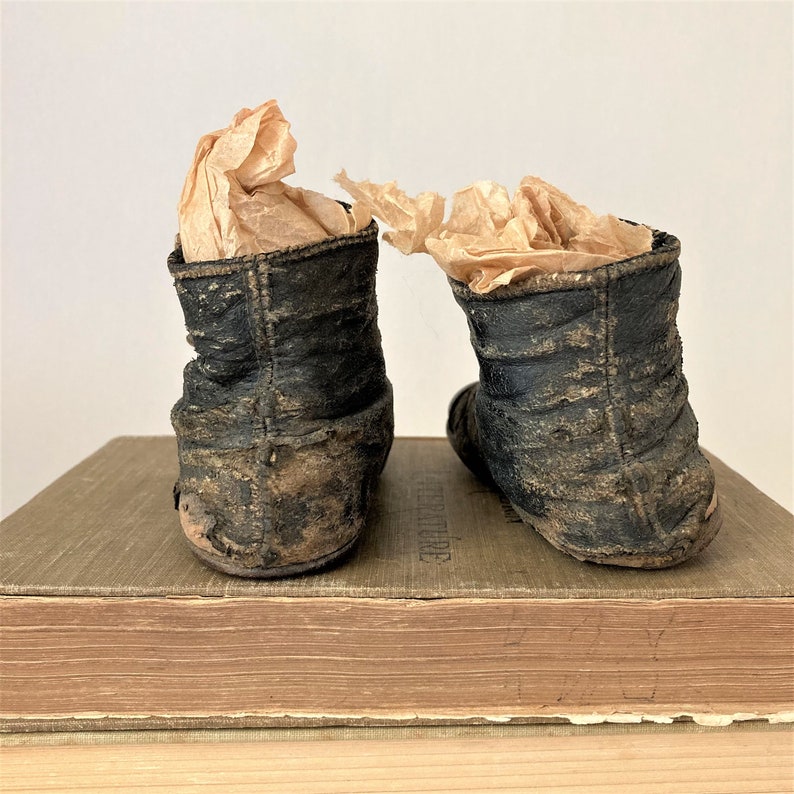 May include: A pair of black leather baby shoes with a worn and distressed look. The shoes are sitting on top of a stack of books.