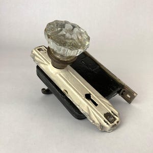 May include: An antique door knob and lock set. The knob is clear glass, and the lock plate is white with a keyhole. The lock is black. The set is likely from the early 20th century.