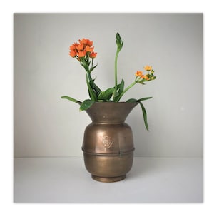 May include: A brass vase with a wide mouth and a heart-shaped emblem that reads "Established 1914" holds a bouquet of orange flowers with green leaves.