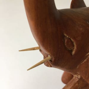 May include: Close-up of a wooden elephant figurine with two toothpicks inserted for tusks. The elephant is carved from reddish-brown wood and has a detailed face.