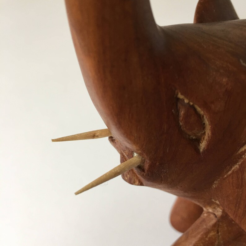 May include: Close-up of a wooden elephant figurine with two toothpicks inserted for tusks. The elephant is carved from reddish-brown wood and has a detailed face.
