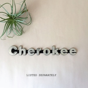 May include: Silver metal letters spelling out the word "Cherokee" on a light pink background. The letters are separated by a black strip.