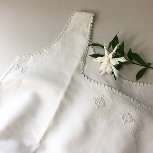 May include: White linen sleeveless top with scalloped edges and embroidered details. The top has the initials "B & L" embroidered on the front.